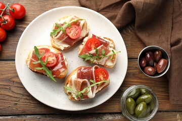 Delicious bruschettas with cream cheese, prosciutto, tomatoes, arugula and olives on wooden table, flat lay