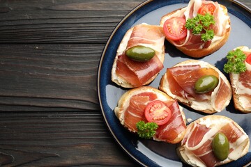 Delicious bruschettas with cream cheese, prosciutto, green olives, tomatoes and parsley on dark wooden table, top view. Space for text