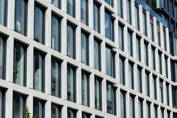 Modern building facade with multiple rectangular windows in grid pattern, featuring glass panes and concrete frames, with greenery at the base during daytime