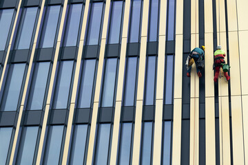 Obraz premium Two workers in safety gear cleaning windows on a high-rise building, suspended by ropes, with vertical glass panels and metal frames surrounding them during daylight