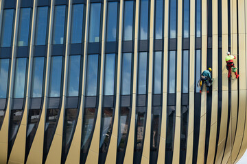 Two workers in safety gear cleaning windows on a modern building facade with vertical stripes and reflective glass panels during daytime with blue sky above