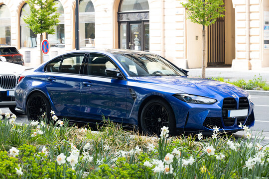 Blue sedan bmw m4 parked on street beside flower bed with white and yellow flowers, green foliage, and trees in background with buildings and signs visible nearby