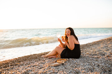 Smiling young woman 35-39 year old holding glass of wine and eating pizza sitting at beach over evening sea outdoors. Summer vacation season.