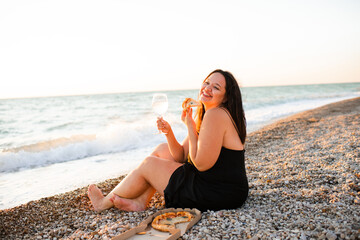 Smiling young woman 35-39 year old holding glass of wine and eating pizza sitting at beach over evening sea outdoors. Summer vacation season.