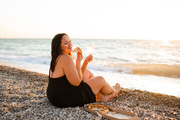 Smiling young woman 35-39 year old holding glass of wine and eating pizza sitting at beach over evening sea outdoors. Summer vacation season.