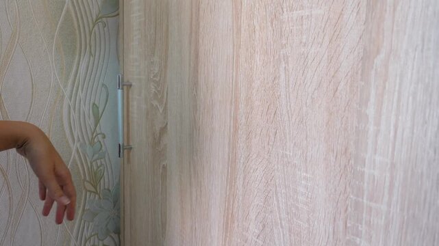 A woman's hand opened and closed the light wooden sliding wardrobe door with a metal handle, revealing a glimpse of clothes arranged on the shelves in the bedroom closet or dressing room