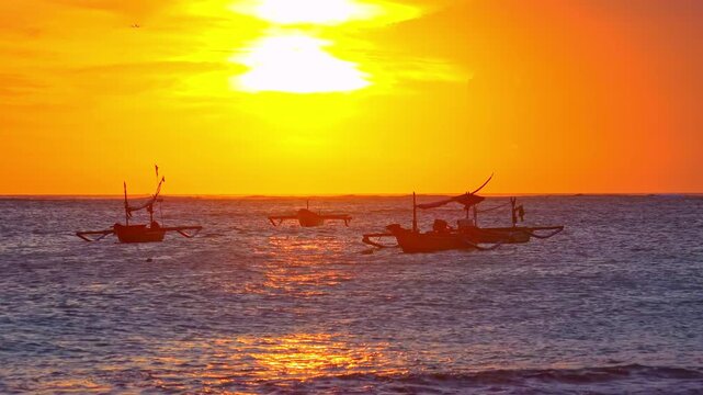 Silhouettes of outrigger fishing boats swaying on sea waves against sun disappearing below horizon. Tropical sunset seascape with ships floating in ocean illuminated by sunlight. Camera zooms out.