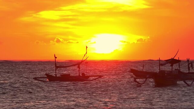 Silhouettes of outrigger fishing boats swaying on sea waves against sun disappearing below horizon. Tropical sunset seascape with ships floating in ocean illuminated by sunlight. Camera zooms out.