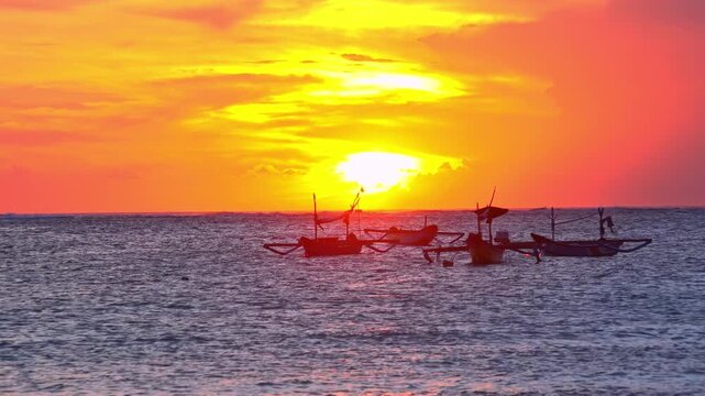 Silhouettes of outrigger fishing boats swaying on sea waves against sun disappearing below horizon. Tropical sunset seascape with ships floating in ocean illuminated by sunlight. Camera zooms out.