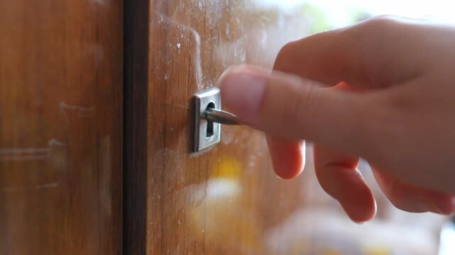 Close-up of a person's hand inserting and turning an old metal key in the keyhole of a vintage wooden cabinet, unlocking or locking the furniture for safety and privacy concepts