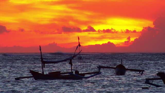 Silhouettes of traditional fishing vessels floating in ocean against red sunset sky with clouds lit by last beams of setting sun on background. Beautiful scene with evening sea. Camera stays still.