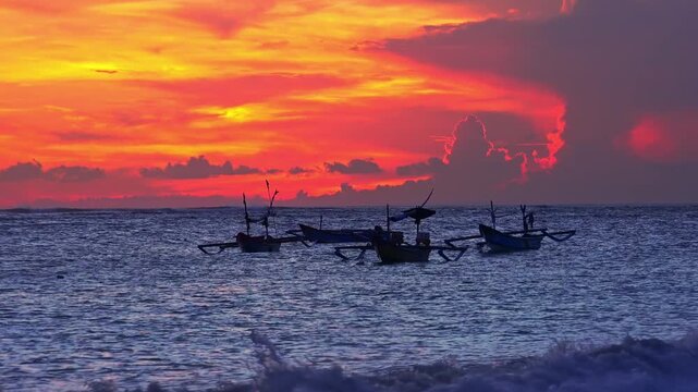 Silhouettes of outrigger fishing boats swaying on sea waves against sun disappearing below horizon. Tropical sunset seascape with ships floating in ocean illuminated by sunlight. Camera zooms out.