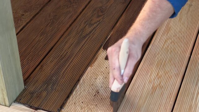 Brown oil for protection of wood by staining. Painting a larch for protecting and pests. Woodworker applying finish to terrace deck board. Repairman varnishing with brush wooden plank floor surface.