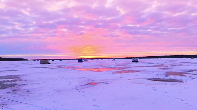 View of a frozen lake with small fishing huts scattered on the ice as the sun sets. The sky shows vibrant colors creating a beautiful sight in winter.