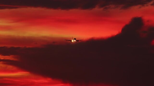 Silhouette of aircraft flying in orange sunset sky with clouds illuminated by setting sun. Gorgeous cloudscape with airplane. Flight of plane with flashing lights at sundown. Camera stays still.