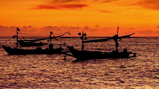 Silhouettes of traditional fishing vessels floating in ocean against red sunset sky with clouds lit by last beams of setting sun on background. Beautiful scene with evening sea. Camera stays still.