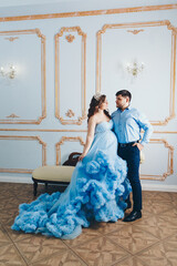 a married couple stands together near a beige sofa. married couple looking at each other. pregnant girl in a lush blue dress. young man in a blue shirt. family photo session. 