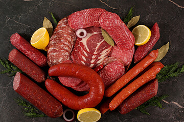 An assortment of various types of sausages, smoked meats, salami, and game sausages with lemon, spices, and herbs on a concrete table. Advertising and banner for a store, menu for a cafe or restaurant