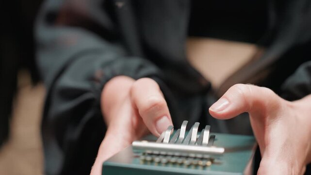 Close up of hands in dark outfit gently plucking metal tines on small green thumb piano, surrounded by soft forest light and tranquil atmosphere, capturing emotion