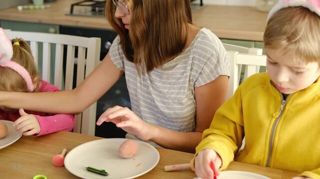 Children siblings painting Easter egg sitting together at table. Easter Family traditions. Teen girl teaching happy little brother and sister to dye, decorate eggs with paints. Preparation to holidays