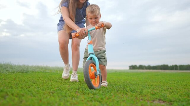 Boy holds handlebar as mother helps with learning. Toddler enjoys outdoor ride. Child learning biking step by step. Mother gently supports child. Boy rides confidently learning and bonding support