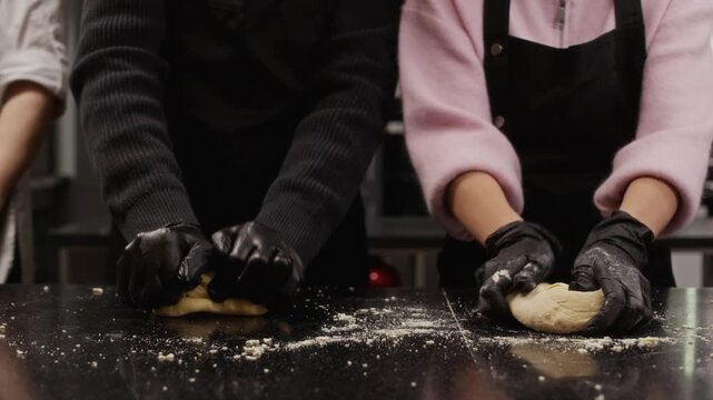 Cropped shot of two culinary course trainees in black gloves kneading dough on countertop while baking pastries in restaurant kitchen during cooking class