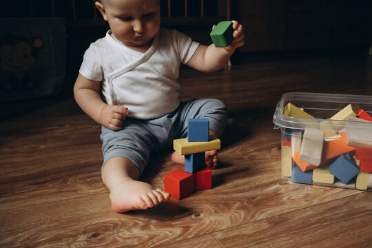 Baby sits on a wooden floor, playfully stacking colorful toy blocks from a nearby container, displaying concentration and curiosity. The scene captures a moment of early development and learning.
