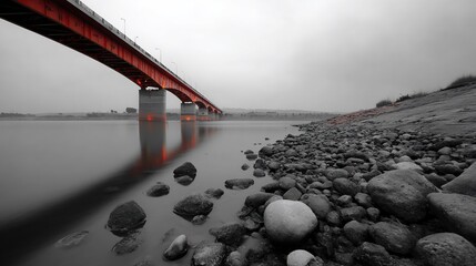 Red bridge spans calm water, rocks lead to shore, grayscale sky above