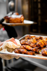 A chef's hands hold a metal baking tray filled with freshly cooked, seasoned chicken wings. Another tray with food is visible in the blurry background, likely in a commercial kitchen or restaurant set
