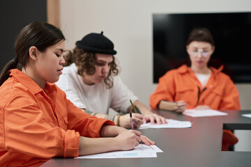 Group of young adults sitting at table completing written assignments in classroom setting, wearing orange prison uniforms, participating in prison education program