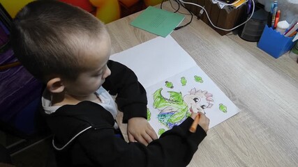 A young child sits at a table using orange and green markers to color in a book. The child smiles and focuses on creating bright images with joy in a sunny room.