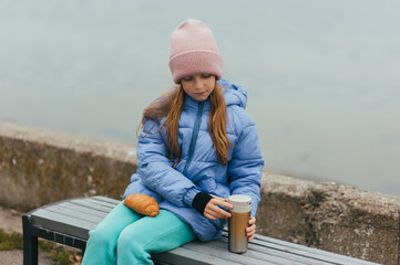 Photograph, portrait of a beautiful teenage girl, child with a thermos and a croissant, sitting on a bench against the backdrop of the sea on a picnic.