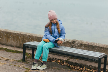 Photograph, portrait of a beautiful teenage girl, child with a thermos of hot tea and croissant, food, sitting on a bench with the sea in the background on a picnic.