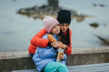 Photo, portrait of a beautiful teenage girl, child with a thermos of hot tea and croissant, food, sitting with her mother, young woman on a bench with the sea in the background on a picnic.