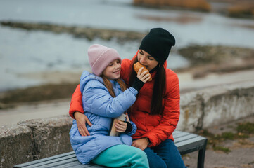 Photo, portrait of teenage girl, child with thermos of hot tea and croissant, food, sitting with mother, young woman on bench against sea background on picnic in autumn.