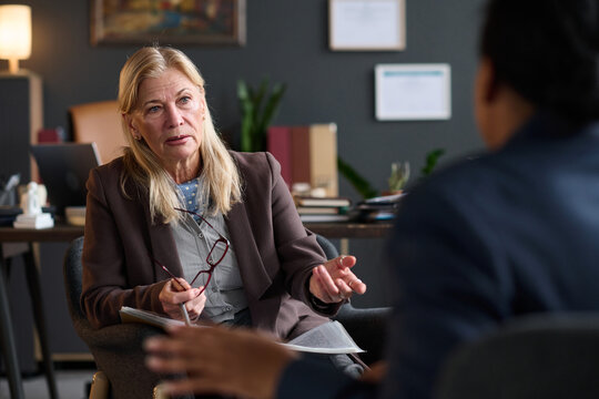 Caucasian therapist listening and gesturing while holding eyeglasses during therapy session with young adult Black woman in office setting, discussing mental health