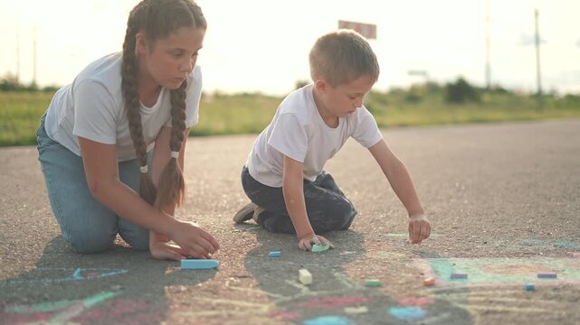 Girl and boy focus on chalk art during summer outdoor play. Children sit on street making creative drawing. Child and kid share joy. Street becomes bright art made by children using colorful chalk.