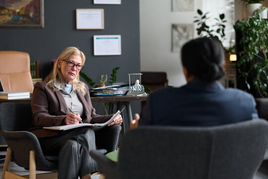Senior Caucasian woman holding clipboard listening attentively to adult woman during therapy session in office setting, both seated facing each other, discussing mental health concerns
