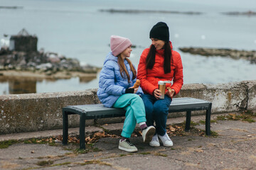 Photo, portrait of a beautiful teenage girl, child with a thermos of hot tea and croissant, food, sitting with her mother, young woman on a bench with the sea in the background on a picnic.