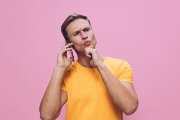 pensive young caucasian man talking phone hand on chin wear orange t-shirt pink background digital...