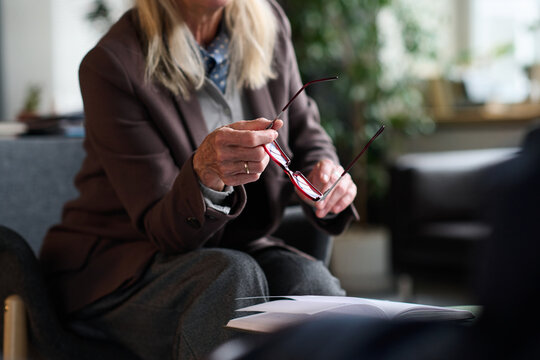 Senior woman holding eyeglasses while sitting with open notebook, engaging in therapy session, only upper body and hands visible, focus on gesture and interaction
