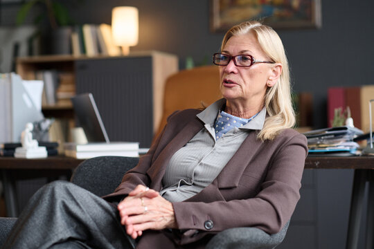 Senior Caucasian woman sitting in office chair talking during therapy session, wearing glasses and business attire, appearing engaged in professional conversation with client