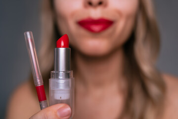 Close-up of red lipstick and lip liner in young blonde woman's hand with blurred lips in...