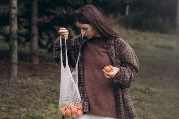 Lifestyle photo of a young woman walking in a forest holding a reusable mesh shopping bag with...