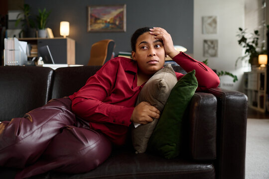 Black young woman reclining on sofa holding cushion, resting head on hand, gazing thoughtfully into distance in modern therapy office setting, appearing contemplative