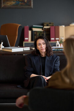 Caucasian woman sitting on sofa attentively listening to client during therapy session in office, bookshelf and desk with folders visible in background