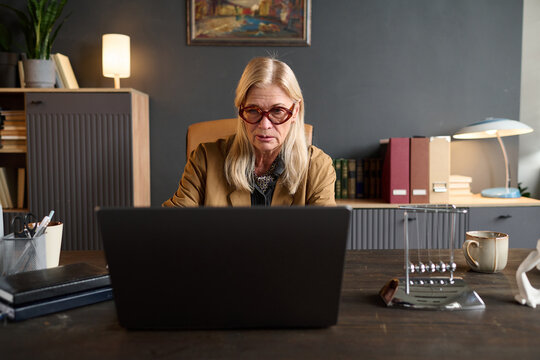 Portrait of senior Caucasian woman wearing glasses sitting at desk using laptop during therapy session, focused expression, office setting with folders and books visible