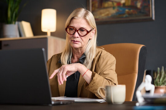 Caucasian senior woman wearing glasses sitting at desk using laptop during online therapy session, focused expression, hand resting near keyboard, modern office background visible