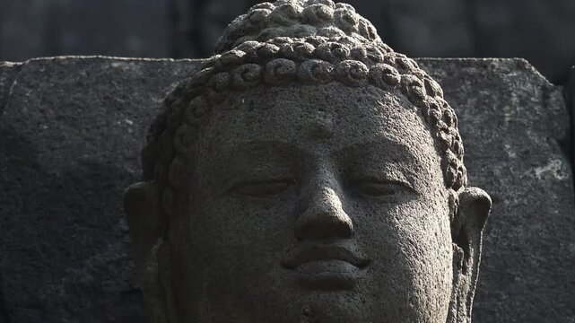 Close-up of head of meditating Buddha's statue carved from dark volcanic rock and illuminated by sun. Sculptural detail of Borobudur Buddhist temple, Magelang, Central Java, Indonesia. Panning video.