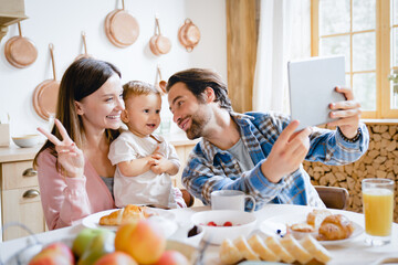 Caucasian young happy family taking selfie photo on tablet, having videocall conversation conference with relatives grandparents at home kitchen. Childcare and parenthood